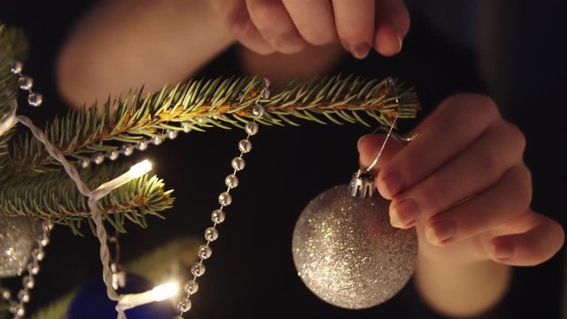 Close-up woman hand is hanging glittery Christmas ball on the branch of Christmas tree. Woman decorating Christmas tree at New year eve