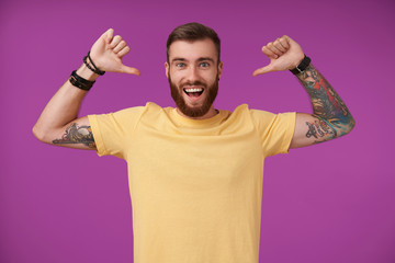 Fototapeta premium Attractive young brunette bearded male with tattooes raising hands and showing on himself with thumbs, smiling widely to camera with self-confidence, standing over purple background in casual clothes