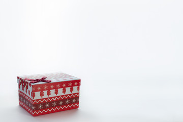 A red Christmas box with gifts and a bow stands on a white background with place for postcard text from the left edge. On the box are Christmas trees and snowflakes.