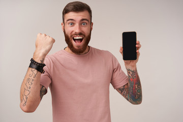 Portrait of joyous tattooed unshaved brunette guy keeping mobile phone in hand and looking at...