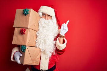 Senior man wearing Santa Claus costume holding tower of gifts over isolated red background very happy pointing with hand and finger to the side