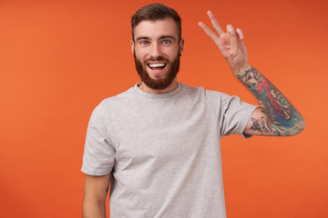 Happy pretty bearded brunette man with tattooes looking at camera cheerfully and smiling broadly, raising fingers with peace sign while standing over orange background