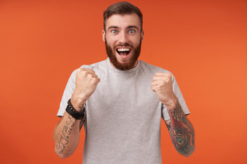 Overjoyed young bearded man with tattooes looking at camera with excited face and keeping raised...