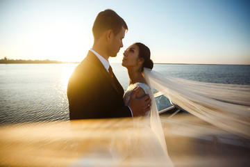 Pretty bride and stylish groom together on the bridge against the background of the boat. Newlyweds enjoy each other tenderly in the shadow of a flying veil. Together. Wedding. Love.