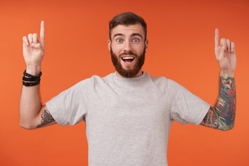 Joyful young tattooed brunette man with beard pointing upwards with raised forefingers and looking at camera with wide eyes and mouth opened, isolated over orange background