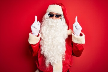 Middle age man wearing Santa Claus costume and sunglasses over isolated red background amazed and surprised looking up and pointing with fingers and raised arms.