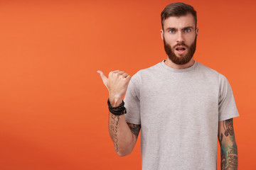 Bewildered tattooed unshaved brunette man with short haircut showing aside with thumb and looking...