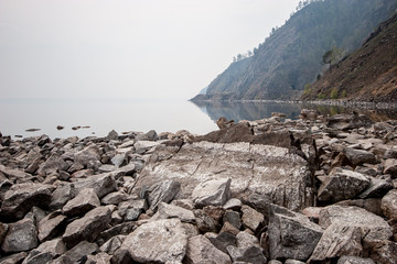 A rock by the lake in fog and large stones by the water. There are trees on the rocks. The stones are gray. Copy space.