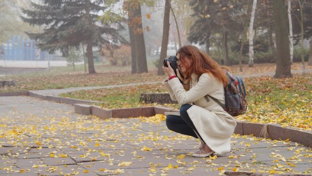 Alone young girl in white coat and backpack taking pictures in a foggy city park. yellow autumn leaves lie around the girl on the ground