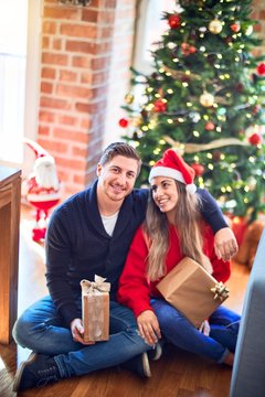 Young beautiful couple smiling happy and confident. Sitting on the floor holding gifts and hugging around christmas tree at home