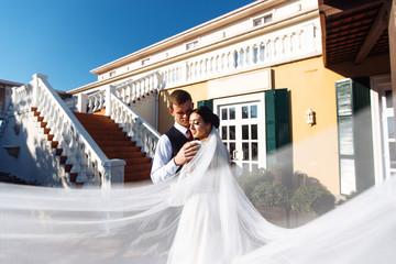 Fototapeta premium Newlyweds enjoy each other tenderly in the shadow of a flying veil. A stylish bridegroom in a black suit gently embraces a beautiful bride in a white dress with a flying veil. Romantic moment.
