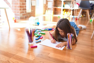 Adorable toddler lying down on the floor drawing using paper and pencils at kindergarten