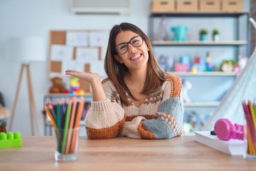 Young beautiful teacher woman wearing sweater and glasses sitting on desk at kindergarten smiling...