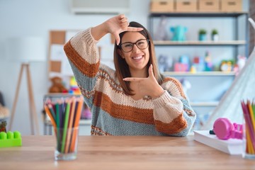 Young beautiful teacher woman wearing sweater and glasses sitting on desk at kindergarten smiling making frame with hands and fingers with happy face. Creativity and photography concept.