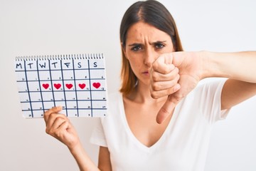 Beautiful redhead woman holding menstruation calendar over isolated background with angry face,...