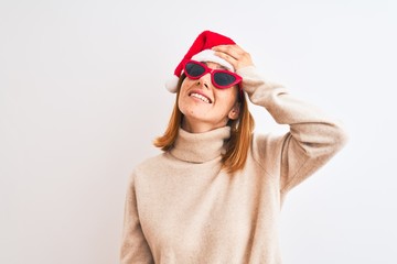 Beautiful redhead woman wearing christmas hat and red sunglasses stressed with hand on head,...