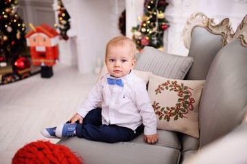 Naklejka premium baby boy in a bright shirt and tie sitting on the sofa on the background of Christmas decor.
