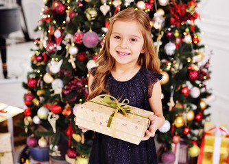 a little cute girl in a smart black dress holding a box with a gift and smiling on the background of the Christmas tree and Christmas decor.