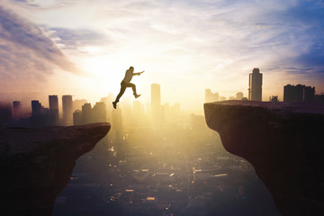 Businessman jumping through gap cliff at sunrise - obrazy, fototapety, plakaty