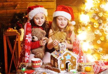 children-two little girls in red Santa hats play with toys covered with a warm blanket, smile, laugh and make a gingerbread house with colored icing on the roof. Christmas vacation.