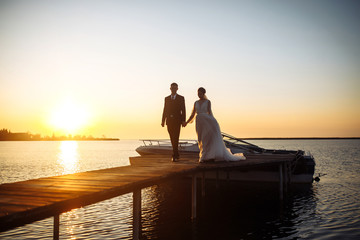Fototapeta premium Beautiful bride and stylish groom together on the bridge against the background of the boat at sunset. Newlyweds tenderly hug, kiss and enjoy each other at sunset. Wedding. Love. Romantic moment. 