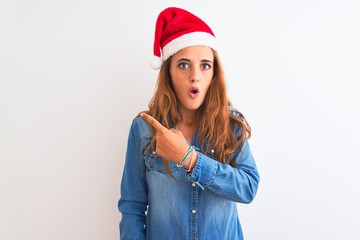 Young beautiful redhead woman wearing christmas hat over isolated background Surprised pointing with finger to the side, open mouth amazed expression.