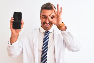 Young scientist man showing smartphone screen over isolated background with happy face smiling doing ok sign with hand on eye looking through fingers