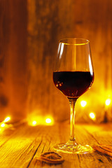 Hot mulled wine (red wine) in a shiny red wine glass on wooden table, decorated with Christmas season spices cinnamon sticks and slice of orange, golden lights
