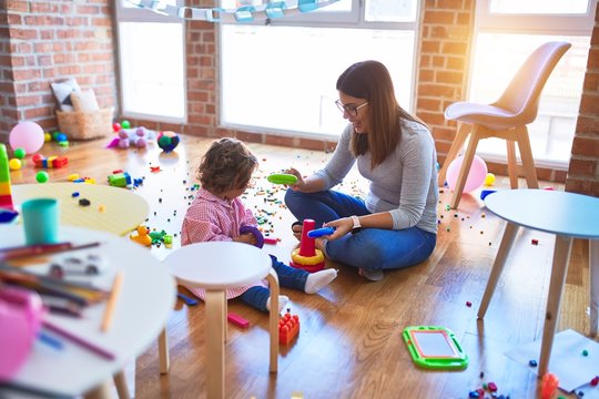 Young beautiful teacher and toddler sitting on the floor building pyramid using hoops at kindergarten