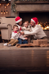 Obraz premium Image of parents in Santa's cap with two sons sitting on floor by fireplace in room
