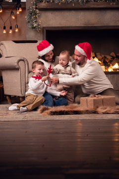 Image of parents in Santa's cap with two sons sitting on floor by fireplace in room