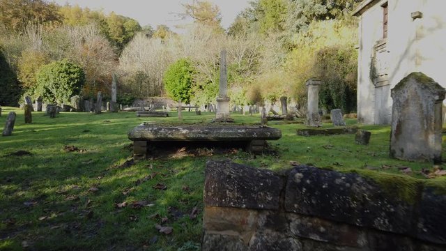 Hand held walk through the entrance gate of a disused rural graveyard between headstones and towards the ruin of an ivy-covered chapel. 
