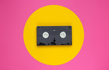 Video cassette on pink background with yellow circle. Studio fashion shot. 80s, pop culture. Top...