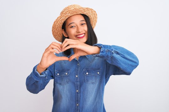 Young beautiful chinese woman wearing denim shirt and hat over isolated white background smiling in love showing heart symbol and shape with hands. Romantic concept.