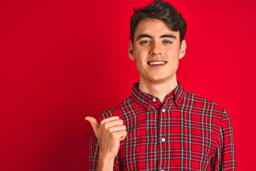 Teenager boy wearing red shirt standing over isolated background smiling with happy face looking and pointing to the side with thumb up.