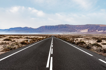 Fototapeta premium long straight empty asphalt road through arid landscape against ocean and mountain range on Lanzarote, Canary Islands