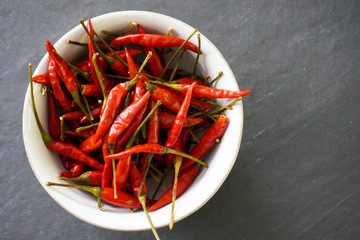 Close-up of a white bowl filled with bright red chiles with green stems on a gray slate background with copy space