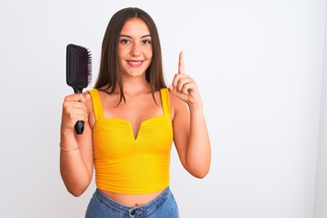 Young beautiful girl holding hair comb standing over isolated white background surprised with an idea or question pointing finger with happy face, number one