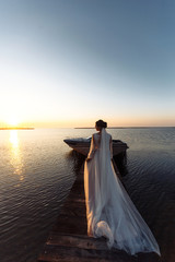 Fototapeta na wymiar Beautiful bride in an elegant white dress on a bridge against the background of the sea and in the rays of sunset. Happy bride with luxury make-up and hairstyle. Wedding day.