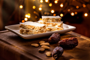 Christmas Concept. Alicante nougat in the foreground, in a warm home atmosphere at Christmas. There are Christmas lights in the background. - obrazy, fototapety, plakaty