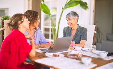 Meeting of middle age women having lunch and drinking coffee. Mature friends smiling happy using laptop at home on a sunny day