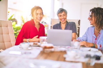Meeting of middle age women having lunch and drinking coffee. Mature friends smiling happy using laptop at home on a sunny day