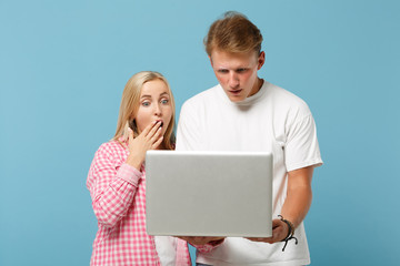 Young shocked couple two friends guy girl in white pink empty blank design t-shirts posing isolated on pastel blue background. People lifestyle concept. Mock up copy space. Holding laptop pc computer.