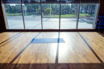 Background of yoga room with yoga mat on wooden floor next to window glass and morning sunlight shine into the room.