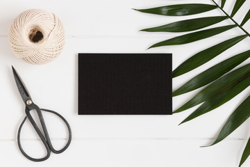 Top view of a black card mockup with workspace accessories and a palm leaf on a white table.