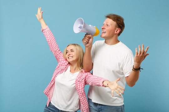 Young happy couple two friends guy girl in white pink empty t-shirts posing isolated on pastel blue wall background. People lifestyle concept. Mock up copy space. Scream in megaphone, spreading hands.