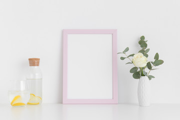 Pink frame mockup with a rose and eucalyptus in a vase, glass and a bottle on a white table. Portrait orientation.