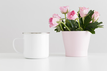 Enamel mug mockup with pink roses in a pot on a white table.