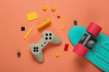 The concept of childhood, entertainment. Cruiser board, gamepad, toy bricks on coral color background. Summer fun. Studio shot. Top view