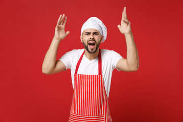 Irritated young bearded male chef cook or baker man in striped apron toque chefs hat posing isolated on red background. Cooking food concept. Mock up copy space. Screaming, swearing, spreading hands.